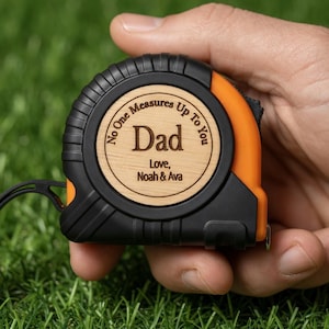 May include: A black and orange tape measure with a wooden center. The wooden center is engraved with the word "Dad", the phrase "No One Measures Up To You", and the names "Love, Noah & Ava". The tape measure is held in a hand.