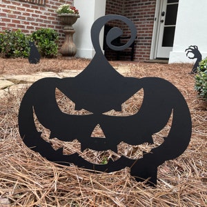 May include: Black metal silhouette of a jack-o'-lantern with a menacing expression. The silhouette is placed in a bed of brown pine straw.