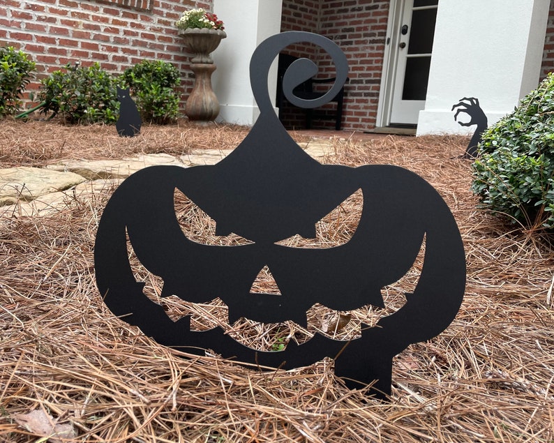 May include: Black metal silhouette of a jack-o'-lantern with a menacing expression. The silhouette is placed in a bed of brown pine straw.