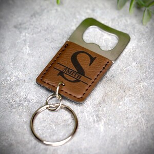 Op de afbeelding: Een bruine leren flessenopener sleutelhanger met een zilveren flessenopener en een gepersonaliseerde initial "S" met de naam "Smith" erop gegraveerd.
