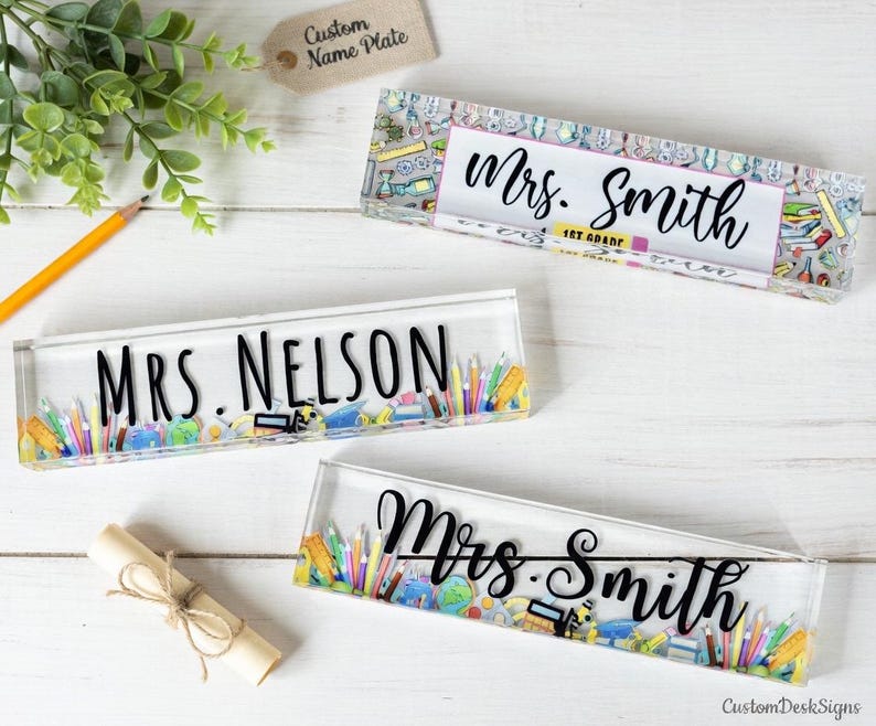 May include: Clear acrylic desk name plates with black lettering. The name plates read "Mrs. Smith" and "Mrs. Nelson". The name plates are decorated with colorful school supplies and the text "1st Grade".