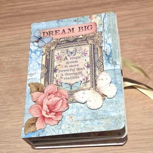 May include: A rectangular notebook with a light blue distressed background featuring a pink rose, two butterflies, and a decorative frame containing the text: DREAM BIG. A single dream is more powerful than a thousand realities. A yellow ribbon is attached.