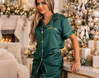 May include: A dark green satin pajama set consisting of a short-sleeved button-down shirt and matching shorts. The shirt features white piping along the collar, front placket, and pockets, with the name Silvia embroidered in gold cursive script.