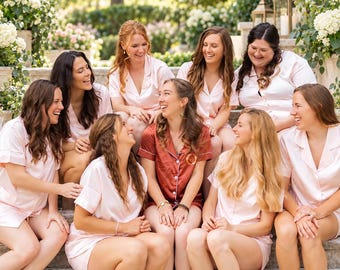 May include: A group of people wearing matching short-sleeved button-down pajama sets. Eight individuals wear light pink satin pajamas, while one individual in the center wears a rust-colored satin pajama set with a small logo on the chest.