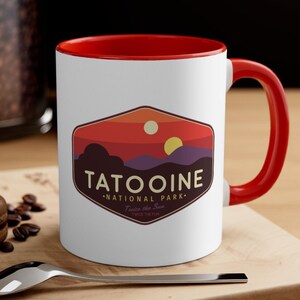 May include: White ceramic mug with a red rim and handle. The mug features a graphic of a sunset with two suns and the text "Tatooine National Park. Twice the Sun. Twice the Fun."