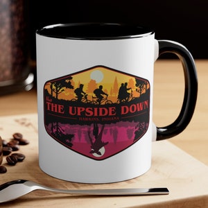 May include: White ceramic mug with a black rim and handle. The mug features a colorful graphic of a retro-style logo with the text "Visit The Upside Down - Hawkins, Indiana" and a silhouette of four figures riding bicycles.