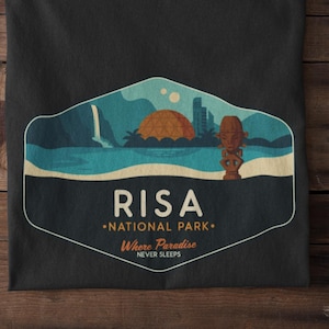 May include: Black t-shirt featuring a graphic of a tropical scene with a waterfall and sunset. The text on the shirt says "RISA NATIONAL PARK Where Paradise Never Sleeps."