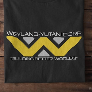 May include: Black t-shirt with the Weyland-Yutani Corp logo in yellow and white. The text "BUILDING BETTER WORLDS" is below the logo. The shirt is folded on a wooden surface, with white sneakers visible.