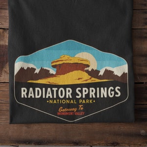 Può includere: Maglietta nera con una grafica in stile vintage di una formazione rocciosa rossa con il testo "RADIATOR SPRINGS NATIONAL PARK. Gateway To MONUMENT VALLEY."