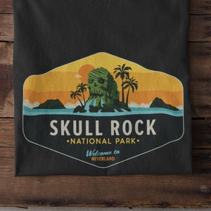 Pode incluir: Camiseta preta com um gráfico de estilo vintage de uma ilha de rocha de crânio com palmeiras. O texto diz "Skull Rock National Park" e "Bem-vindo a Neverland".
