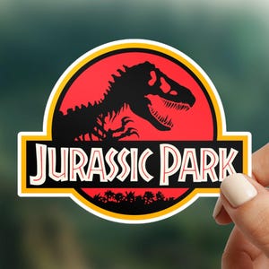 Könnte beinhalten: Ein Aufkleber mit dem Jurassic Park-Logo. Er zeigt eine schwarze Tyrannosaurus Rex-Silhouette auf rotem Hintergrund, mit den Worten "JURASSIC PARK" in Weiß auf einem schwarzen Rechteck, das gelb umrandet ist. Ein klassisches Film-Emblem.