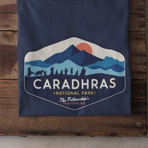 Puede incluir: Una camiseta azul marino con un gráfico de una cordillera y el texto "Caradhras National Park - The Fellowship's Frostbitten Trail".