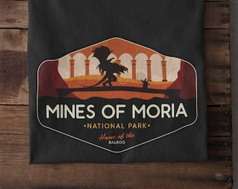 Op de afbeelding: Zwart t-shirt met een afbeelding van een draak, bogen en de tekst "MINES OF MORIA - NATIONAL PARK - Home of the BALROG". Het shirt is opgevouwen op een houten oppervlak, met witte sneakers zichtbaar.