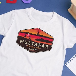 May include: White t-shirt featuring a graphic of a red, orange, and yellow sunset with two figures fighting with lightsabers. The graphic is in a hexagon shape with the text "Mustafar - National Park - Sand in the air" in black.