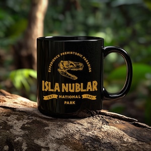 May include: Black ceramic mug with a gold graphic featuring a dinosaur skull and the text "ISLA NUBLAR PARK". The mug also includes the text "EXPERIENCE PREHISTORIC PARADISE" and "EST. NATIONAL 1993". The mug is set against a blurred green background.