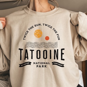 May include: A beige sweatshirt with a graphic design featuring the text "Tatooine National Park" and the words "Twice the sun, twice the fun" in a circle around the text. The graphic also includes a stylized image of two suns and a wavy line.