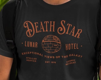 Puede incluir: Una camiseta negra con un gráfico naranja de la Estrella de la Muerte de Star Wars. El gráfico dice "Death Star - Lunar Hotel - Exceptional Views of the Galaxy - Stellar Service Est. 1976 Cosmic Retreat"