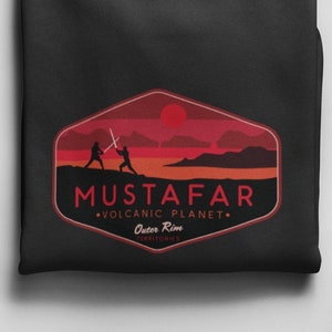 Peut inclure: Sweat-shirt noir avec un graphique rouge et orange d'une planète avec deux personnages se battant avec des sabres laser. Le texte "Mustafar - Planète volcanique - Territoires de la Bordure Extérieure" est imprimé sous le graphique.