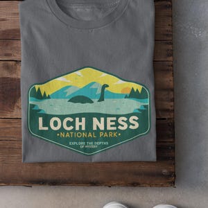 May include: A gray t-shirt with a green and yellow graphic of a Loch Ness Monster in a lake. The graphic says "Loch Ness National Park" and "Explore the Depths of Mystery".