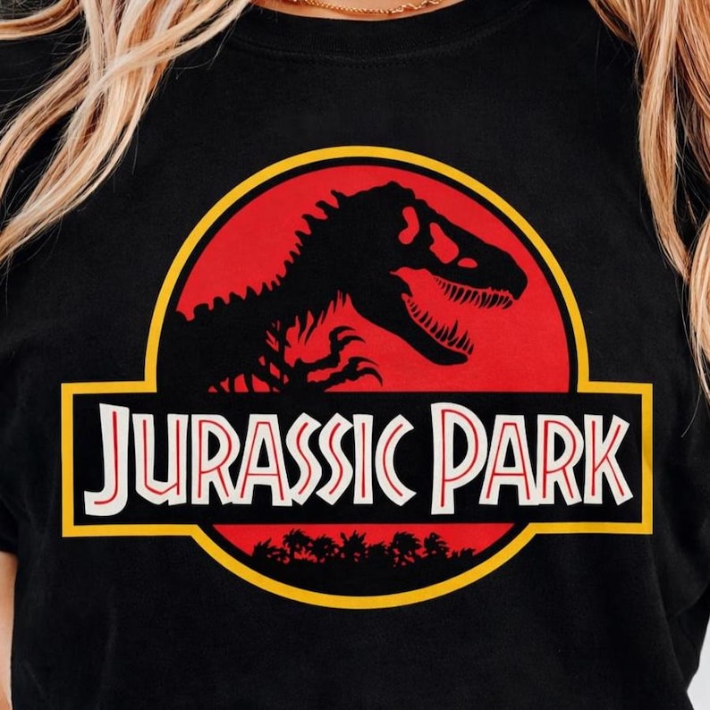 May include: Black t-shirt featuring the Jurassic Park logo. The design includes a red circle with a black dinosaur silhouette, a yellow border, and the words "JURASSIC PARK" in white.