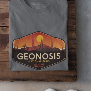 May include: A grey t-shirt featuring a vintage-style graphic of the fictional planet Geonosis from Star Wars. The graphic depicts a sunset over a rocky landscape with the text "Geonosis National Park. Witness the Great Battle."