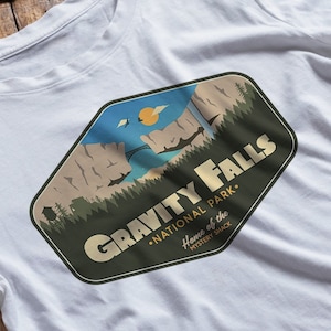 K&ouml;nnte beinhalten: Ein wei&szlig;es T-Shirt mit einem gr&uuml;nen, braunen und blauen Grafikdruck eines Gebirges mit einer Br&uuml;cke und dem Text "Gravity Falls National Park. Home of the Mystery Shack."