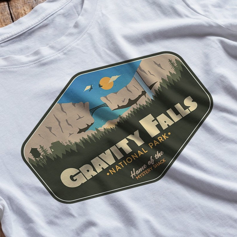 K&ouml;nnte beinhalten: Ein wei&szlig;es T-Shirt mit einem gr&uuml;nen, braunen und blauen Grafikdruck eines Gebirges mit einer Br&uuml;cke und dem Text "Gravity Falls National Park. Home of the Mystery Shack."