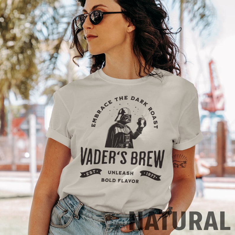 May include: A white t-shirt with a black and white graphic of Darth Vader holding a cup of coffee. The graphic includes the text "Embrace the Dark Roast" and "Vader's Brew". The text below the graphic reads "Est. 1977" and "Unleash Bold Flavor".