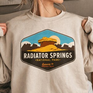 以下が含まれることがあります： 「RADIATOR SPRINGS NATIONAL PARK」のテキストが入った、砂漠の風景のカラフルなグラフィックが特徴のライトベージュのスウェットシャツ。 デザインには岩の形成、山、太陽が含まれています。 スウェットシャツはクルーネックです。