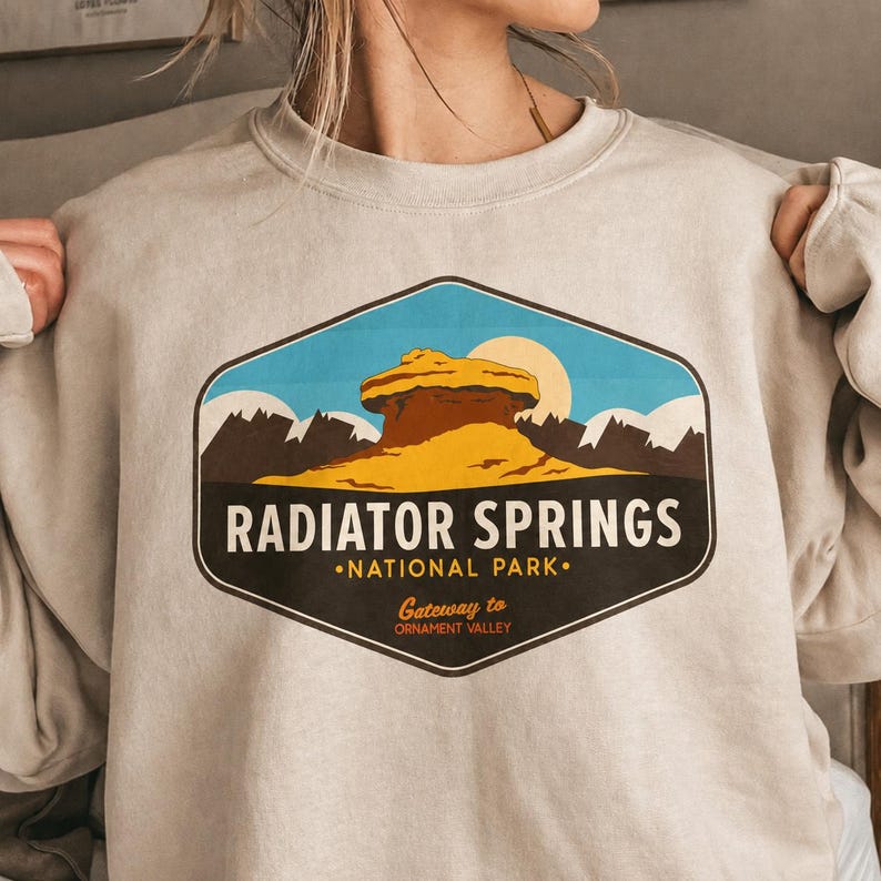 以下が含まれることがあります： 「RADIATOR SPRINGS NATIONAL PARK」のテキストが入った、砂漠の風景のカラフルなグラフィックが特徴のライトベージュのスウェットシャツ。 デザインには岩の形成、山、太陽が含まれています。 スウェットシャツはクルーネックです。