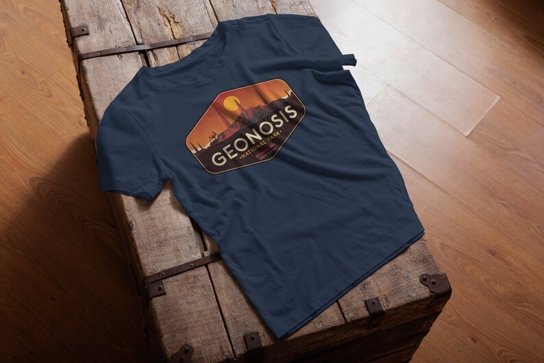 May include: A navy blue t-shirt featuring a vintage-style logo for Geonosis National Park. The logo depicts a sunset scene with a cityscape and the text "Geonosis National Park".
