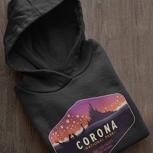 Puede incluir: Sudadera con capucha negra con un dise&ntilde;o gr&aacute;fico. El dise&ntilde;o presenta el texto "CORONA NATIONAL PARK" y una ilustraci&oacute;n de paisaje con linternas flotantes. El dise&ntilde;o incluye las frases "SEE THE LIGHTS" y "ENJOY THE SIGHTS".