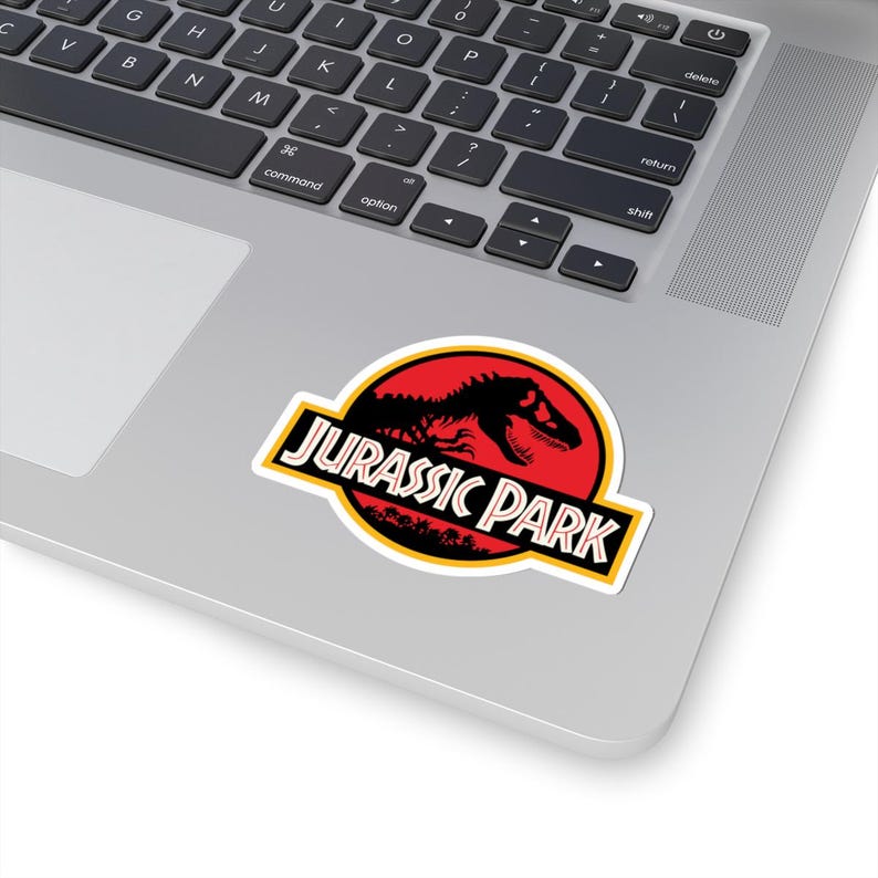 May include: A sticker featuring the "Jurassic Park" logo. The design includes a red circle with a black dinosaur silhouette, the words "JURASSIC PARK" in a yellow-bordered black rectangle, and a green tree line.