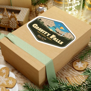 May include: A brown cardboard gift box with a green ribbon. The box has a sticker on it that says "Gravity Falls National Park - Home of the Mystery Shack".