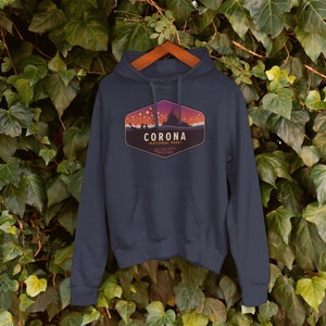 Puede incluir: Sudadera con capucha azul marino con un gr&aacute;fico frontal. El gr&aacute;fico presenta la palabra "CORONA" sobre "NATIONAL PARK" en un dise&ntilde;o estilizado con una silueta de castillo y cielo estrellado. La sudadera est&aacute; colgada en una percha de madera.