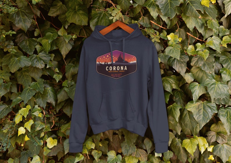 Puede incluir: Sudadera con capucha azul marino con un gr&aacute;fico frontal. El gr&aacute;fico presenta la palabra "CORONA" sobre "NATIONAL PARK" en un dise&ntilde;o estilizado con una silueta de castillo y cielo estrellado. La sudadera est&aacute; colgada en una percha de madera.