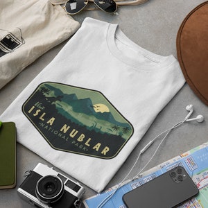 K&ouml;nnte beinhalten: Ein wei&szlig;es T-Shirt mit einem gr&uuml;n-braunen Vintage-Logo, auf dem "Besuchen Sie Isla Nublar - Nationalpark" steht. Das Logo zeigt eine Bergkette, Palmen und eine Dinosauriersilhouette.