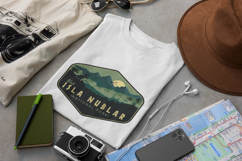 K&ouml;nnte beinhalten: Ein wei&szlig;es T-Shirt mit einem gr&uuml;n-braunen Vintage-Logo, auf dem "Besuchen Sie Isla Nublar - Nationalpark" steht. Das Logo zeigt eine Bergkette, Palmen und eine Dinosauriersilhouette.
