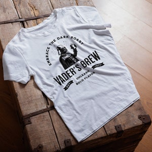 May include: White t-shirt with a black graphic of Darth Vader holding a cup of coffee. The graphic says "Embrace the Dark Roast, Vader's Brew, Unleash Bold Flavor".