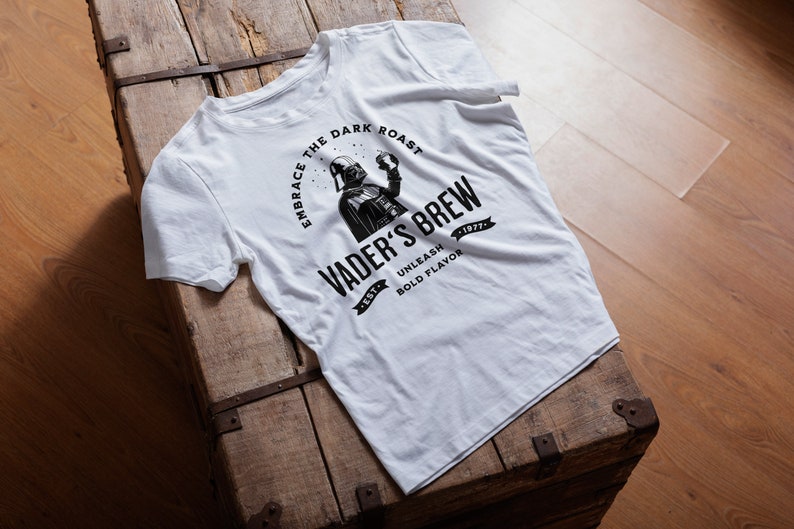 May include: White t-shirt with a black graphic of Darth Vader holding a cup of coffee. The graphic says "Embrace the Dark Roast, Vader's Brew, Unleash Bold Flavor".