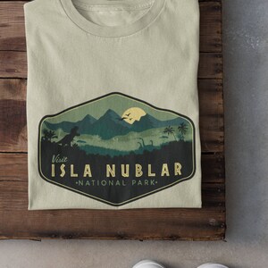 K&ouml;nnte beinhalten: Ein hellgr&uuml;nes T-Shirt mit einer Grafik im Vintage-Stil, die einen Dinosaurier und eine Bergkette zeigt. Die Grafik lautet "Besuchen Sie Isla Nublar National Park."