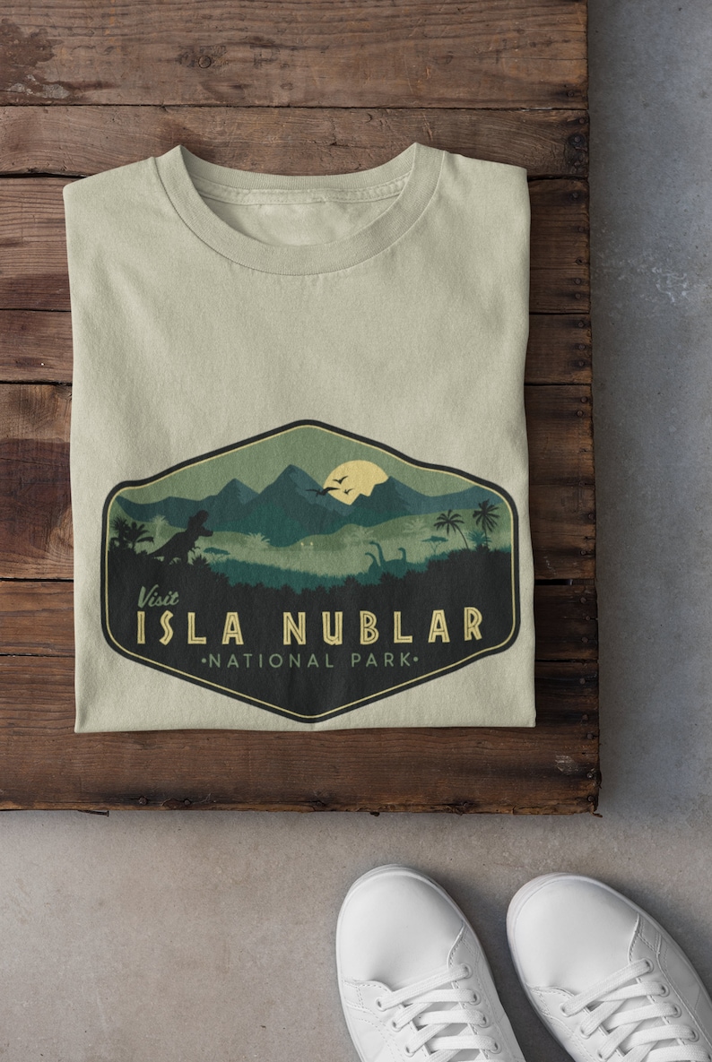 K&ouml;nnte beinhalten: Ein hellgr&uuml;nes T-Shirt mit einer Grafik im Vintage-Stil, die einen Dinosaurier und eine Bergkette zeigt. Die Grafik lautet "Besuchen Sie Isla Nublar National Park."
