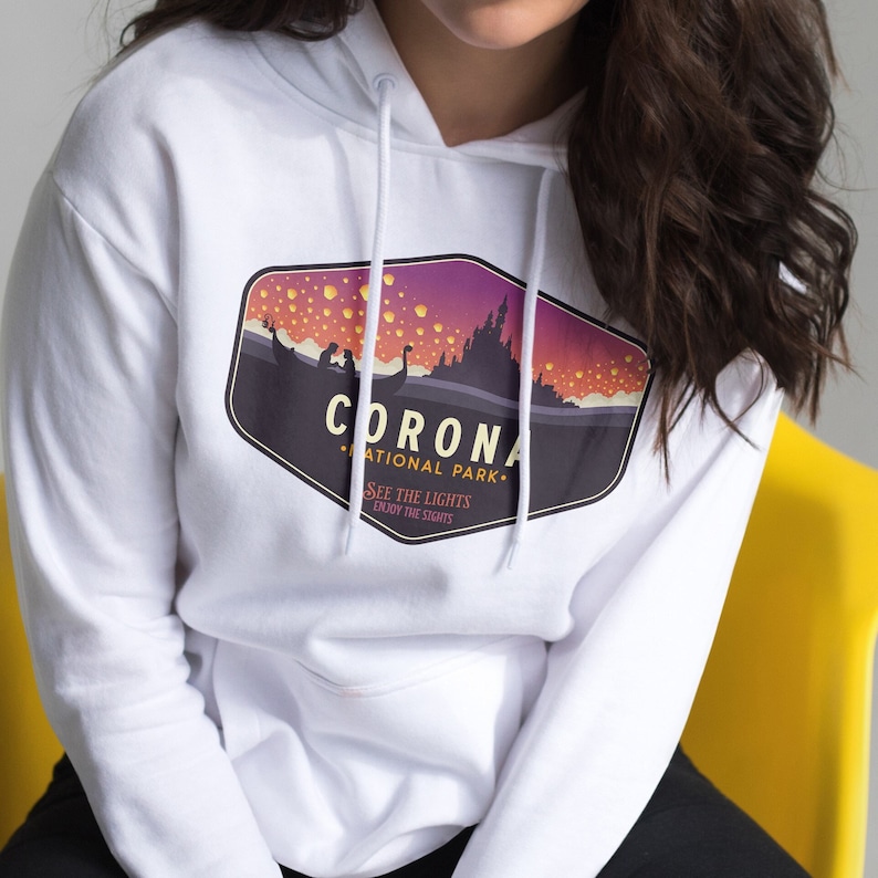 Puede incluir: Sudadera con capucha blanca con un dise&ntilde;o gr&aacute;fico de una escena nocturna con el texto "CORONA NATIONAL PARK". El dise&ntilde;o incluye una silueta de castillo, un barco y linternas flotantes. Tambi&eacute;n incluye el texto "SEE THE LIGHTS, ENJOY THE SIGHTS".