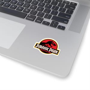 May include: A sticker featuring the Jurassic Park logo. The design includes a red circle with a black dinosaur silhouette and the words "JURASSIC PARK" in a yellow and black rectangular border. The sticker is on a laptop.