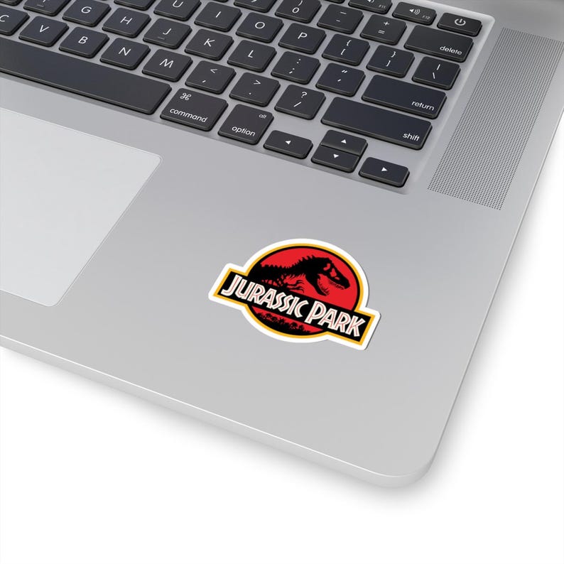 May include: A sticker featuring the Jurassic Park logo. The design includes a red circle with a black dinosaur silhouette and the words "JURASSIC PARK" in a yellow and black rectangular border. The sticker is on a laptop.