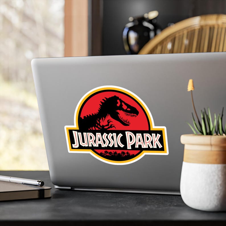 May include: A sticker featuring the Jurassic Park logo. The design includes a black dinosaur silhouette inside a red circle, with the words "JURASSIC PARK" in a yellow and white rectangular banner below. The sticker is on a laptop.
