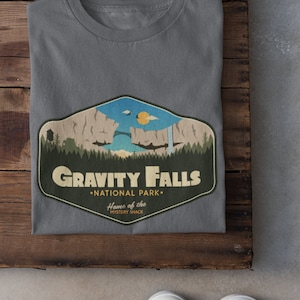 K&ouml;nnte beinhalten: Graues T-Shirt mit einem Grafikdruck eines Retro-Nationalpark-Logos f&uuml;r Gravity Falls, einer fiktiven Stadt aus der Zeichentrickserie "Gravity Falls". Das Logo zeigt eine Bergkette, eine Br&uuml;cke, einen Wasserfall und den Text "Gravity Falls National Park" und "Home of the Mystery Shack".