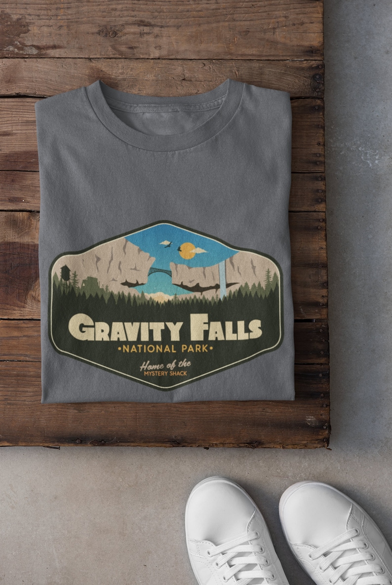 K&ouml;nnte beinhalten: Graues T-Shirt mit einem Grafikdruck eines Retro-Nationalpark-Logos f&uuml;r Gravity Falls, einer fiktiven Stadt aus der Zeichentrickserie "Gravity Falls". Das Logo zeigt eine Bergkette, eine Br&uuml;cke, einen Wasserfall und den Text "Gravity Falls National Park" und "Home of the Mystery Shack".