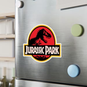 May include: A rectangular magnet featuring the "Jurassic Park" logo. The design includes a black Tyrannosaurus Rex silhouette against a red background, with the words "JURASSIC PARK" in white letters. A yellow border surrounds the logo.