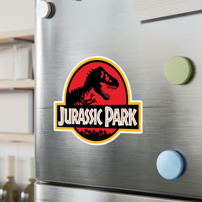 May include: A rectangular magnet featuring the "Jurassic Park" logo. The design includes a black Tyrannosaurus Rex silhouette against a red background, with the words "JURASSIC PARK" in white letters. A yellow border surrounds the logo.